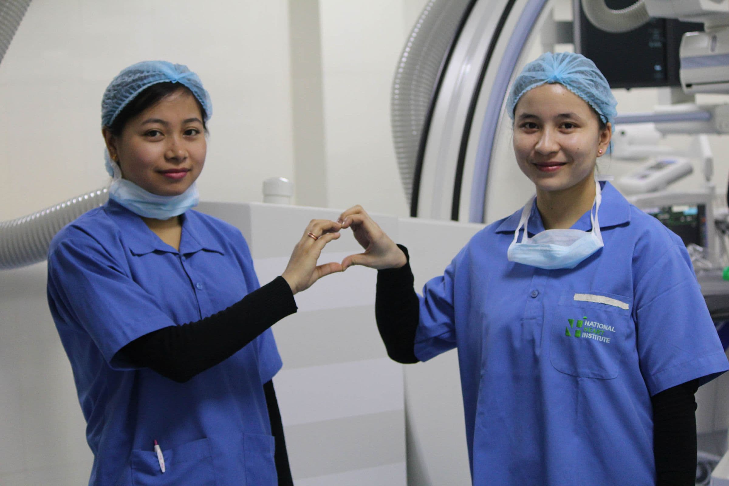 Two National Heart Institute staff members in scrubs making a heart sign in the cath lab